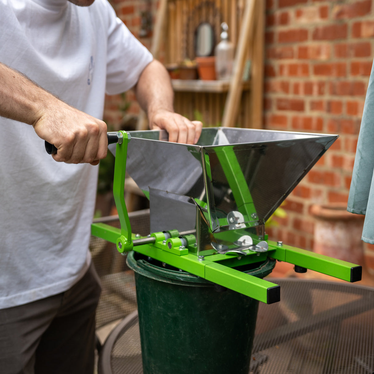 Draagbare fruitmolen met slinger in glanzend roestvrij staal en groene handgreep, geplaatst boven een groene emmer voor fruitverwerking