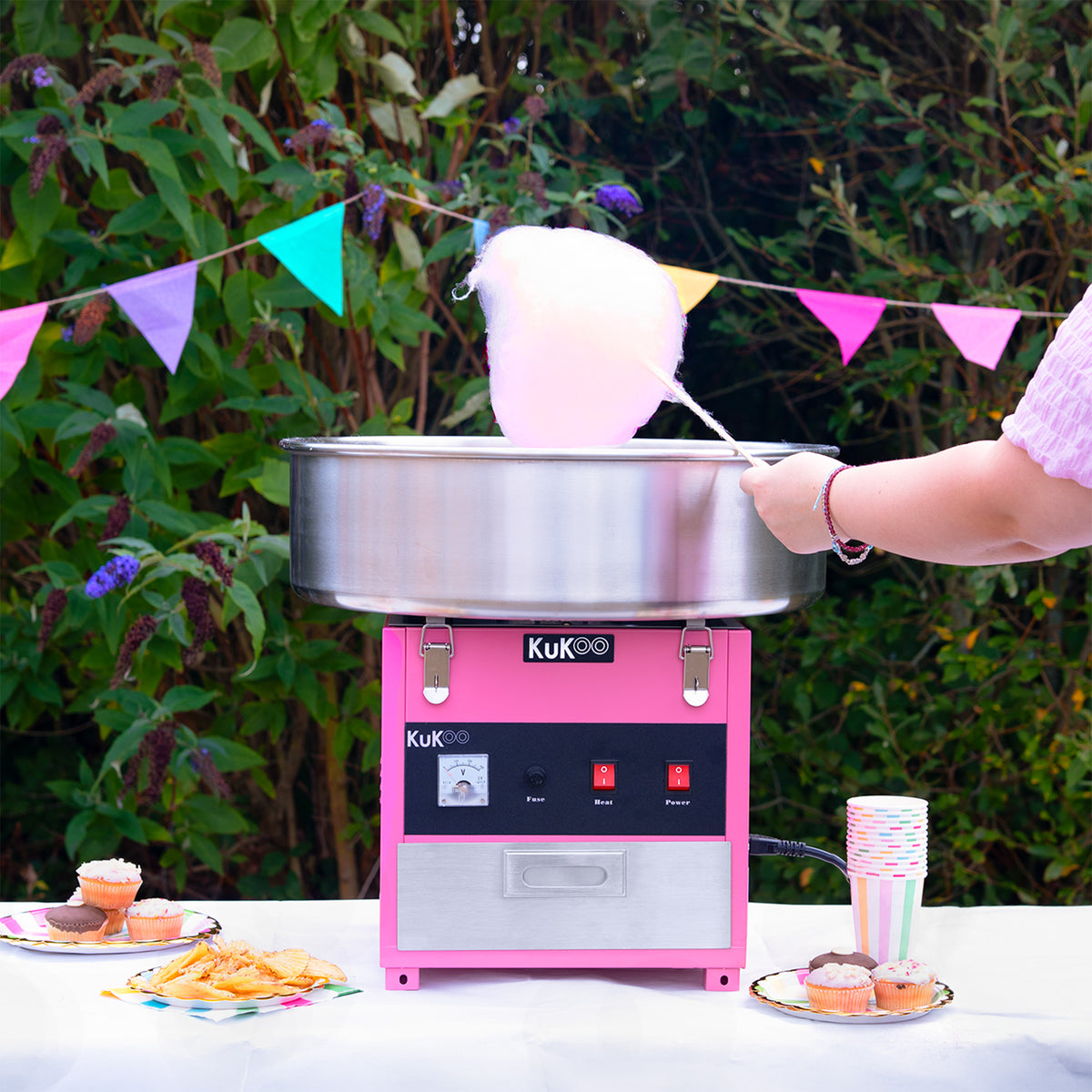 Roze suikerspinmachine met een glanzende metalen kom, omringd door kleurrijke feestdecoraties en lekkernijen op een tafel