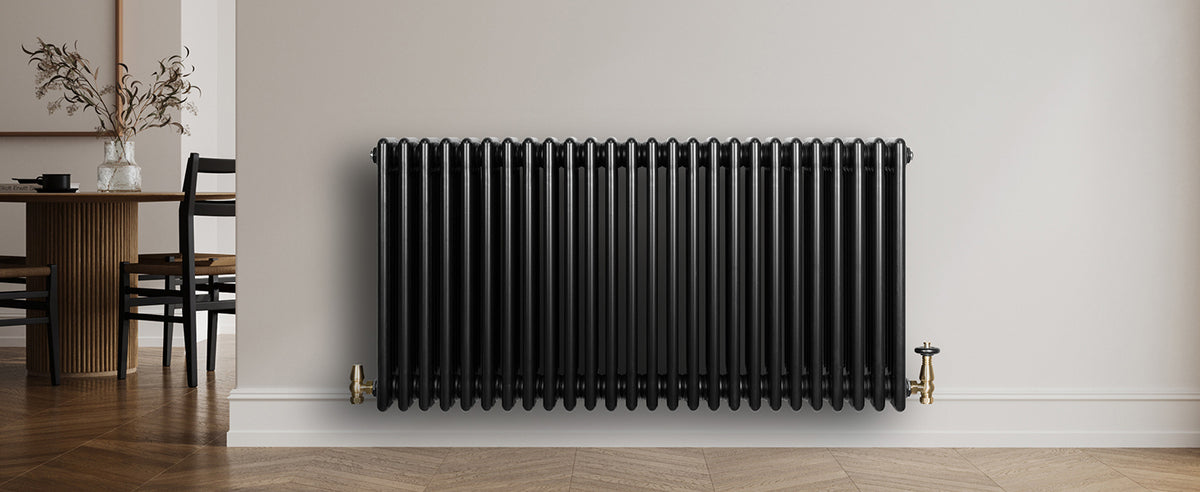 Zwart gietijzeren radiator met verticale ribben, geplaatst tegen een lichte muur, met gouden aansluitingen en een houten vloer op de achtergrond