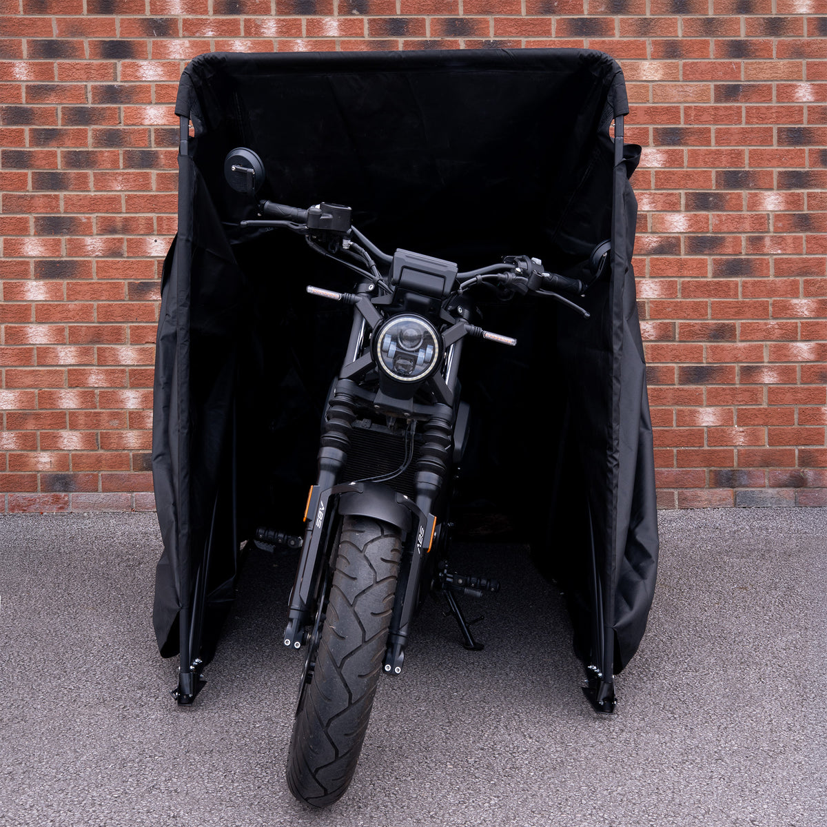 Zwarte motorfiets met een robuuste band, gedeeltelijk verborgen in een zwarte hoes tegen een bakstenen muur