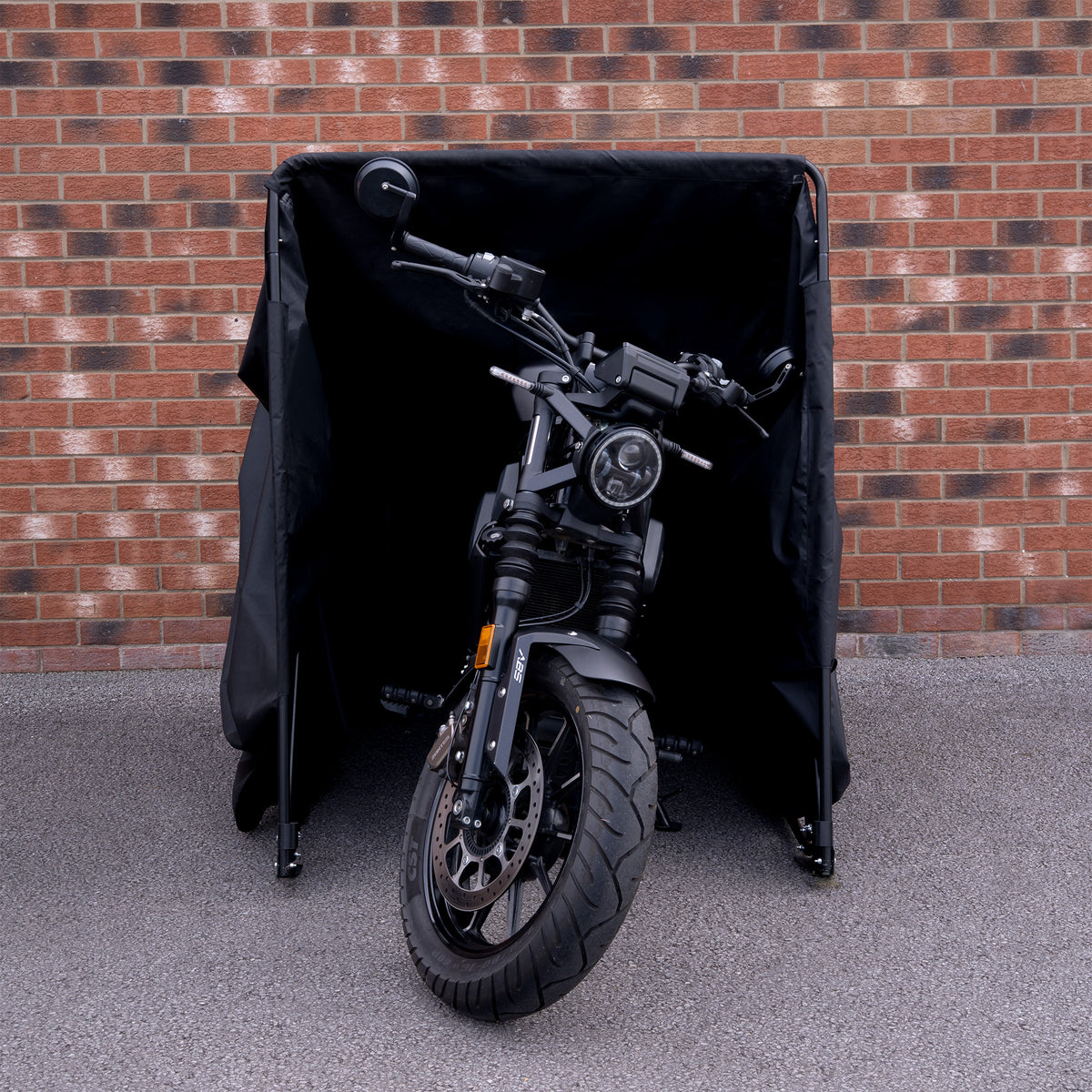 Zwarte motorfiets met een robuuste uitstraling, gedeeltelijk verborgen in een beschermende zwarte hoes tegen een bakstenen muur