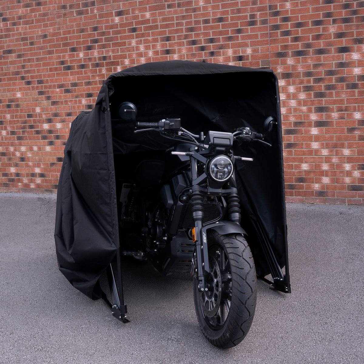 Zwarte motorfiets gedeeltelijk bedekt met een waterdichte hoes, met een moderne uitstraling en opvallende koplampen, tegen een bakstenen muur