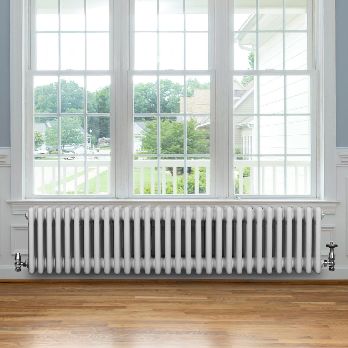 Traditionele witte 4-kolom radiator van 300 x 1370 mm met haakse thermostaatkraan, geplaatst onder een groot raam met uitzicht op de tuin