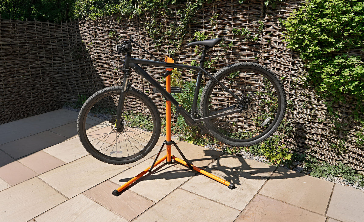 Zwart mountainbike op een oranje fietsondersteuner, met een groene achtergrond van planten en een stenen vloer in de tuin