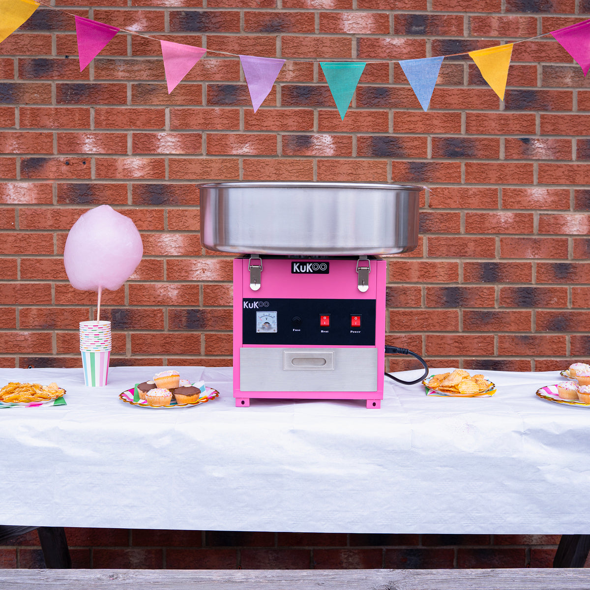 Roze KuKoo suikerspinmachine met roestvrijstalen kom, omringd door kleurrijke vlaggetjes en zoete lekkernijen op een feesttafel