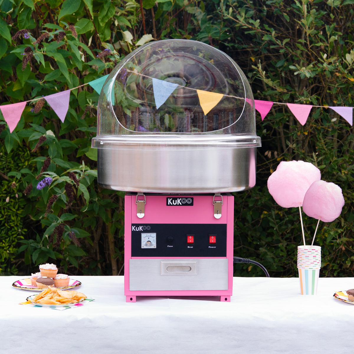 Suikerspinmachine in roze met een glanzende metalen kom, omringd door kleurrijke vlaggetjes en zoete lekkernijen op een tafel