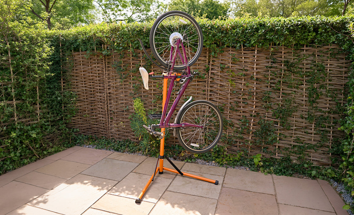 Een paarse fiets op een oranje fietsstandaard, met een groene achtergrond van hagen en een betegelde ondergrond in de tuin