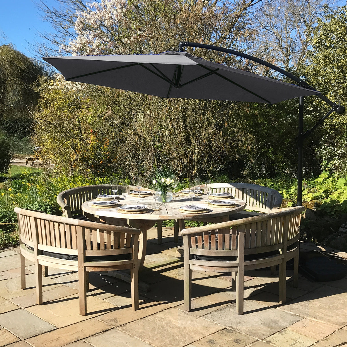Zweefparasol in grijs boven een houten tafel met zes stoelen, omringd door groene planten en bloemen in een zonnige tuin