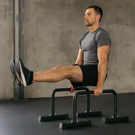 Een man in een grijs T-shirt en zwarte shorts voert een oefening uit op een paar stevige zwarte dip bars in een moderne gymomgeving