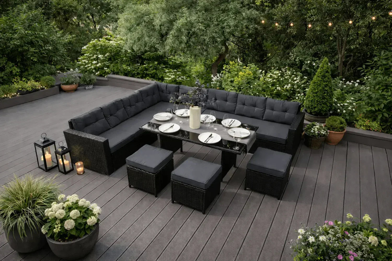 Moderne tuinset met een zwarte, zachte bank, glazen tafel en sfeervolle verlichting, omringd door groene planten en bloemen