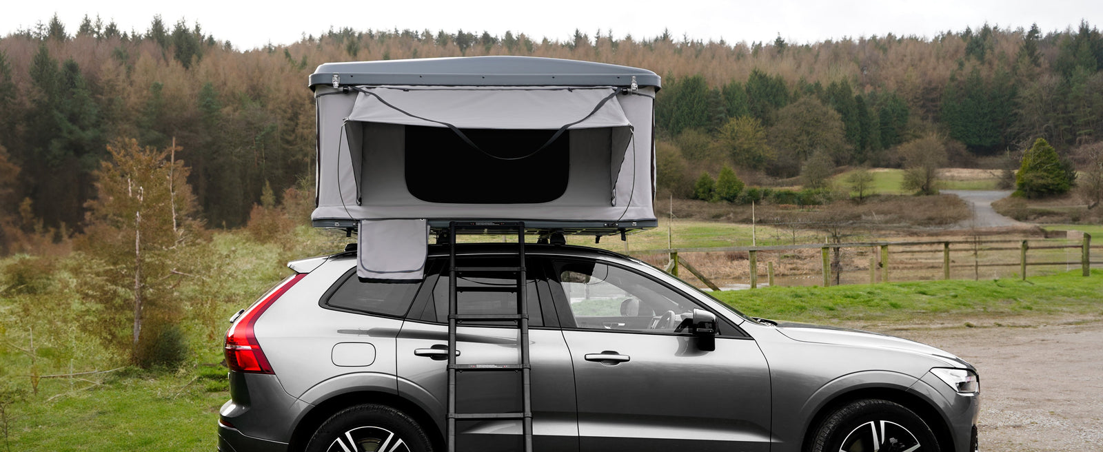 Een grijze daktent op een SUV, met een stevige ladder, omringd door groene bossen en een open veld, perfect voor kampeerliefhebbers