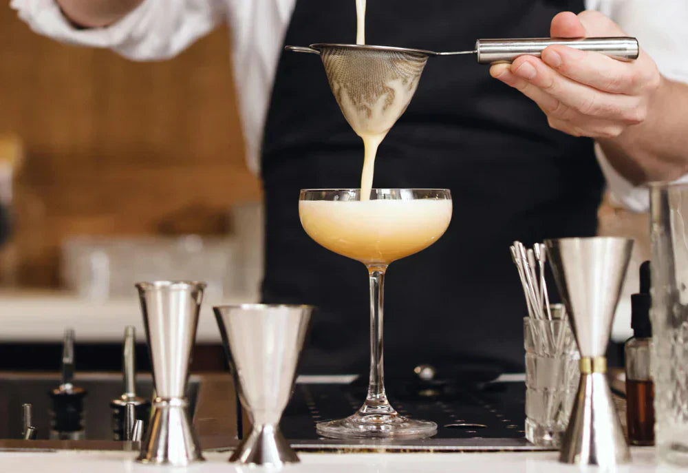 Een barman giet een romige cocktail door een zeef in een elegante coupe, omringd door jiggers en baraccessoires