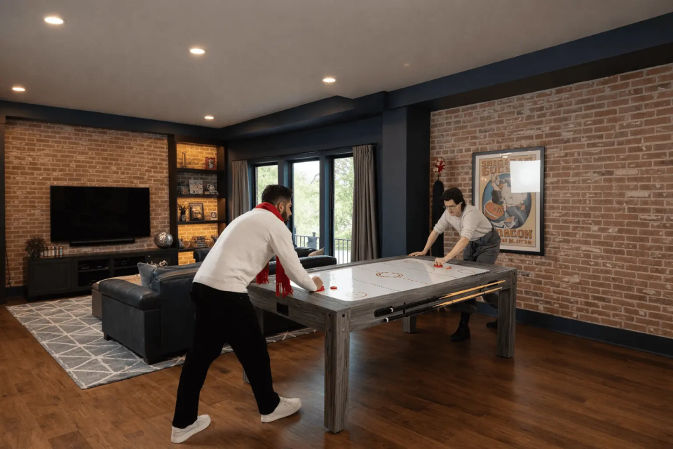Twee mensen spelen aan een moderne 7-in-1 spellentafel in een stijlvolle woonkamer met bakstenen muur en houten vloer