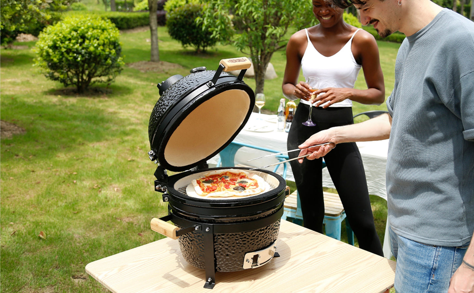 Een Kamado BBQ met een pizza erop, omringd door groene tuin, terwijl twee mensen genieten van een drankje en koken in de buitenlucht