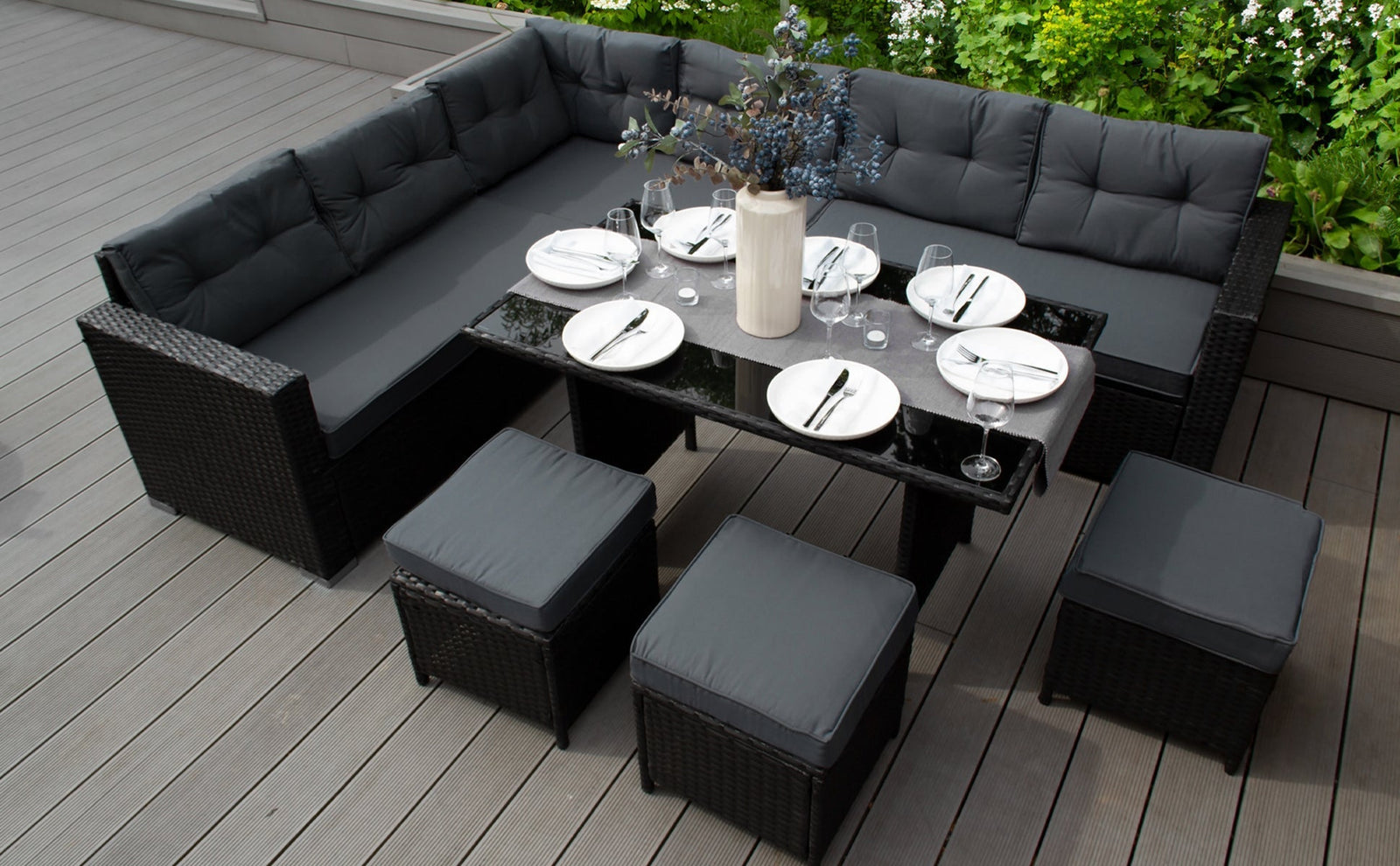 Moderne rotan tuinset met grijze kussens, een zwart glazen tafelblad en stijlvolle stoelen, omringd door groene planten