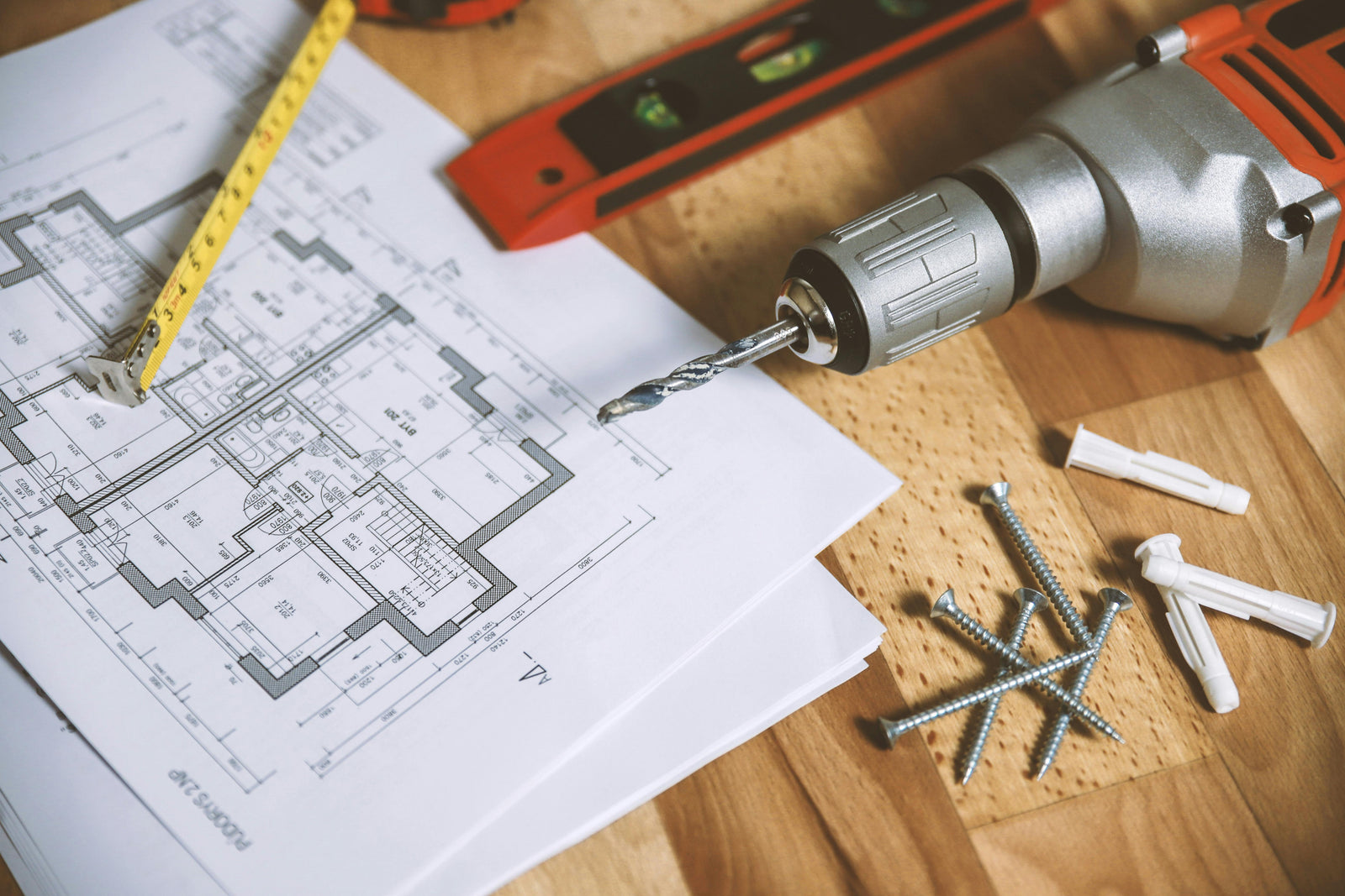 Een plattegrond van een woning ligt op een houten tafel, omringd door een boormachine, schroeven, pluggen en een meetlint