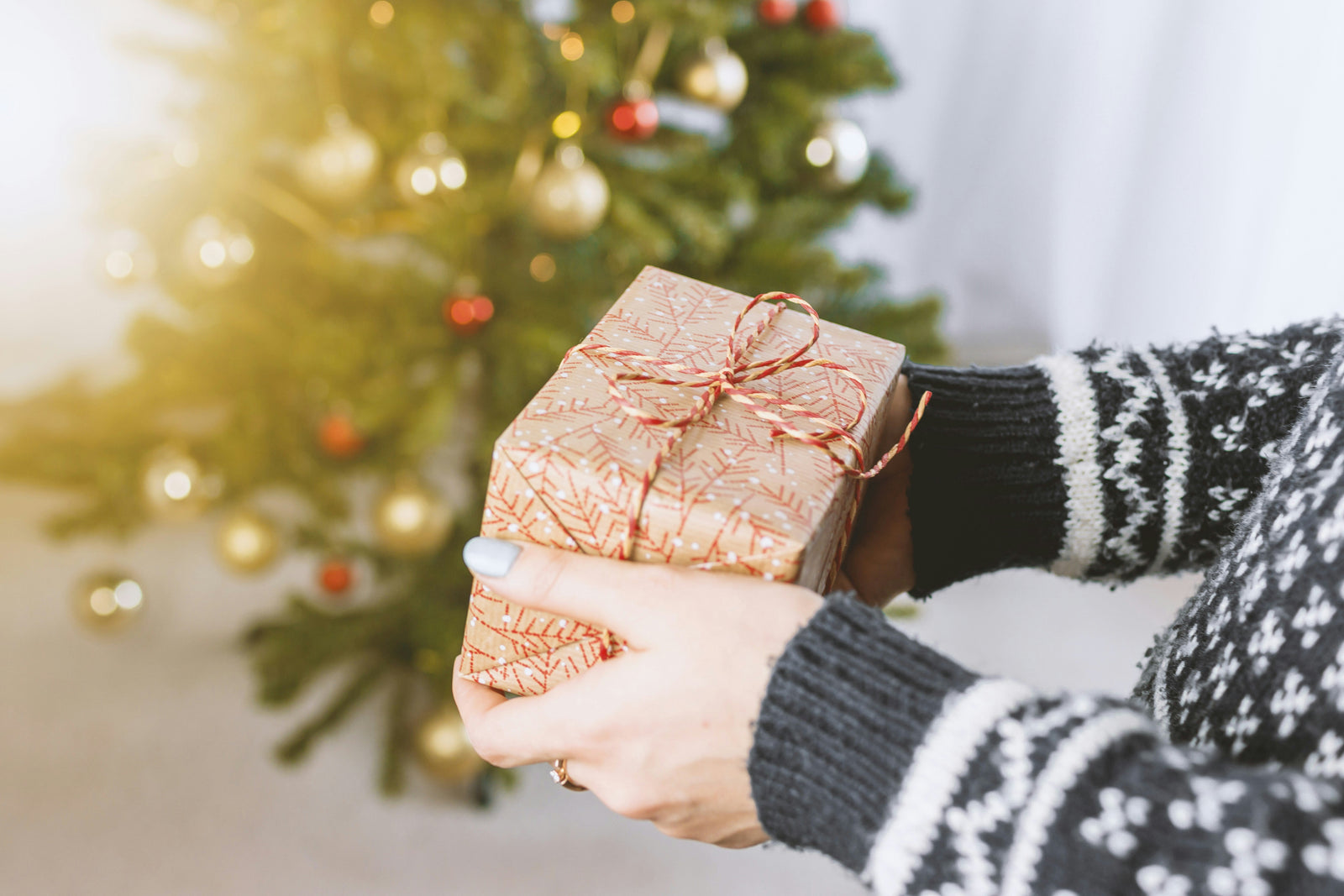 Een persoon in een gebreide trui houdt een mooi ingepakt cadeau met een gouden strik vast, voor een kerstboom met versieringen