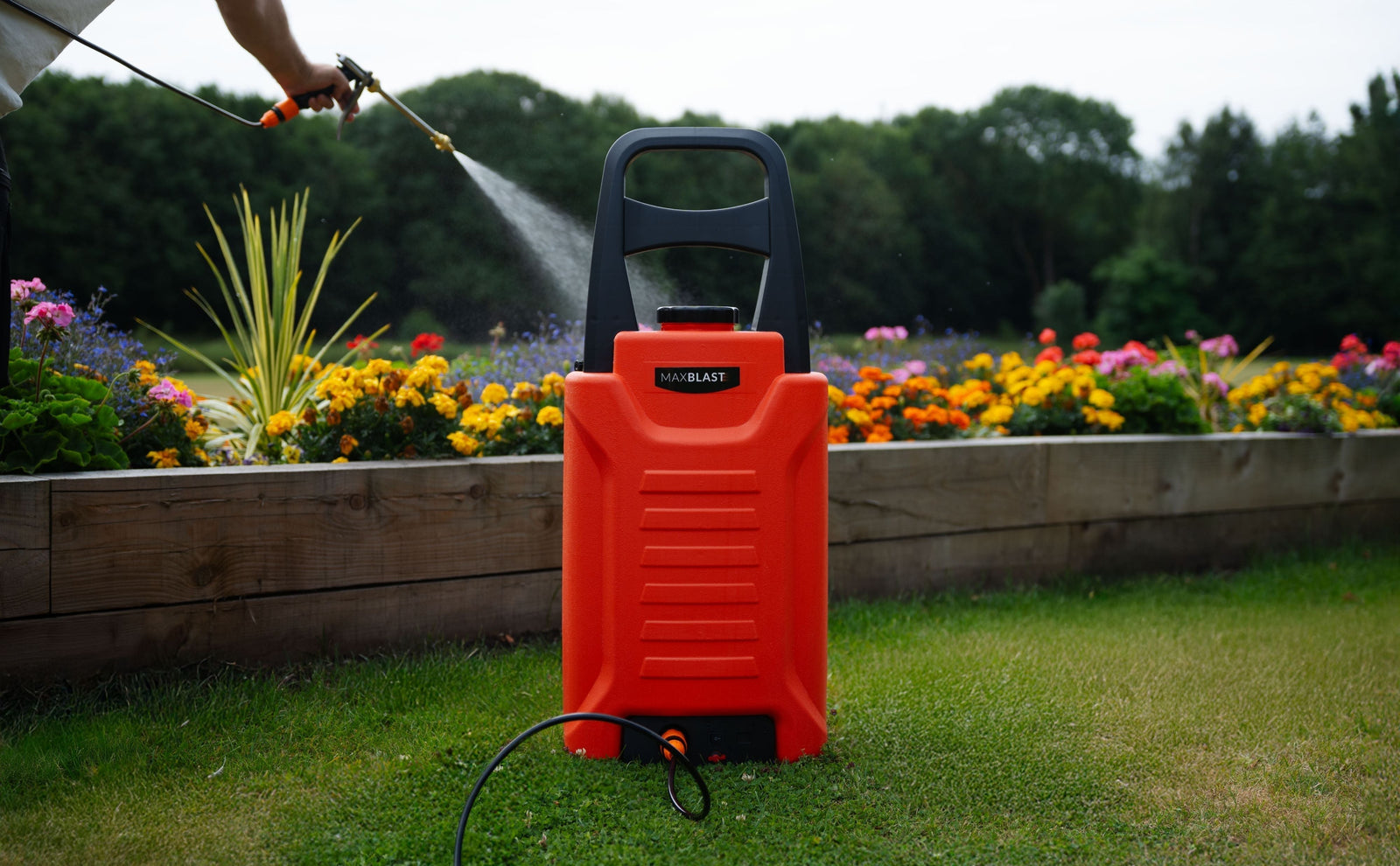Heldere oranje 30L watertank op wieltjes, gevuld met water, naast kleurrijke bloemen in een tuin, met een persoon die sproeit