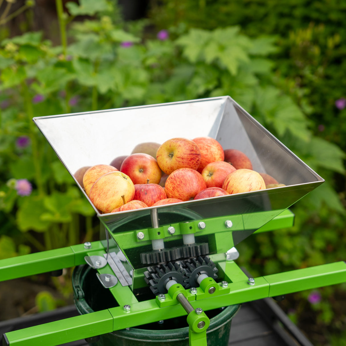 Draagbare fruitmolen met slinger, groene metalen structuur, roestvrijstalen kom vol met rode appels, ideaal voor fruitverwerking