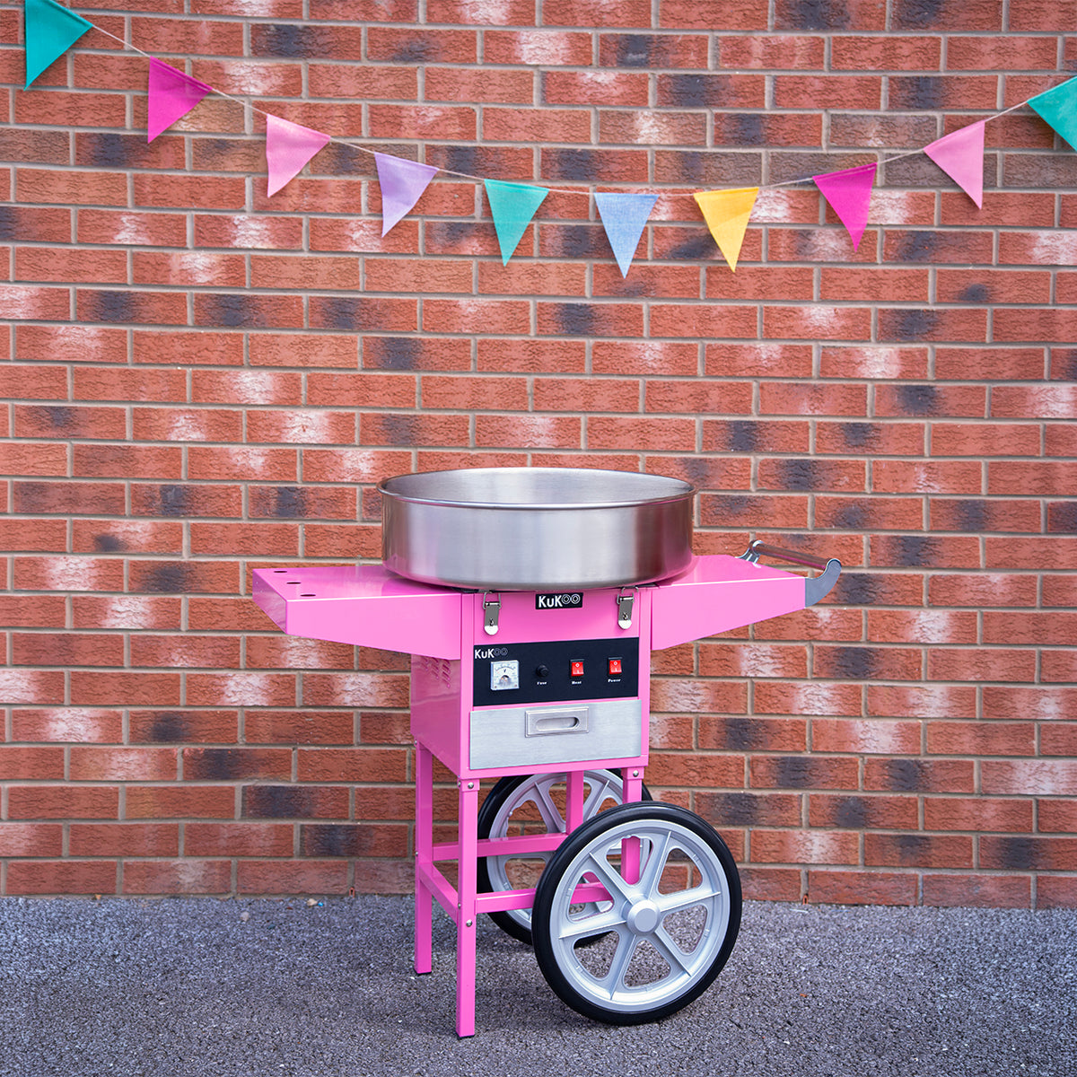 Roze suikerspinmachine met een glanzende metalen kom, geplaatst voor een bakstenen muur versierd met kleurrijke vlaggetjes
