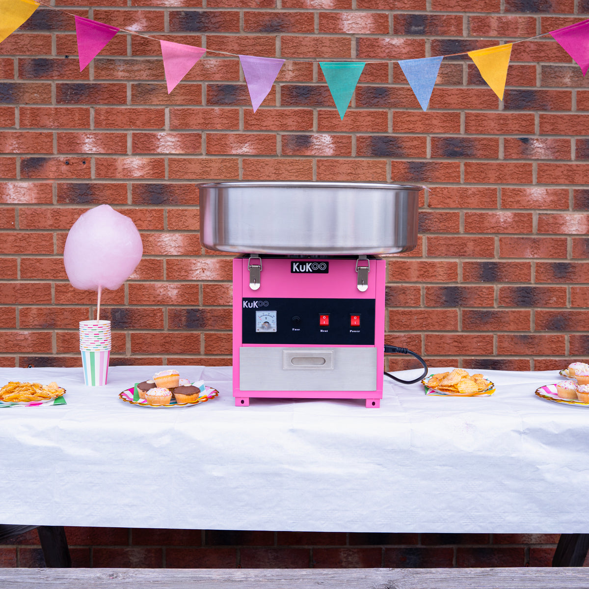 Roze suikerspinmachine op een tafel met een witte tafelkleed, versierd met kleurrijke vlaggetjes en lekkernijen zoals cupcakes en suikerspin