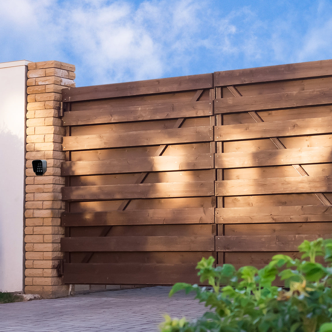 Houten poort met moderne lijnen, omgeven door een stenen pilaar en groene planten, onder een heldere blauwe lucht