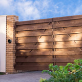 Houten poort met moderne lijnen, omgeven door een stenen pilaar en groene planten, onder een heldere blauwe lucht