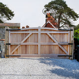 T-Mech swing poort met houten planken, metalen scharnieren en een grindpad, omgeven door groene bomen en een huis op de achtergrond