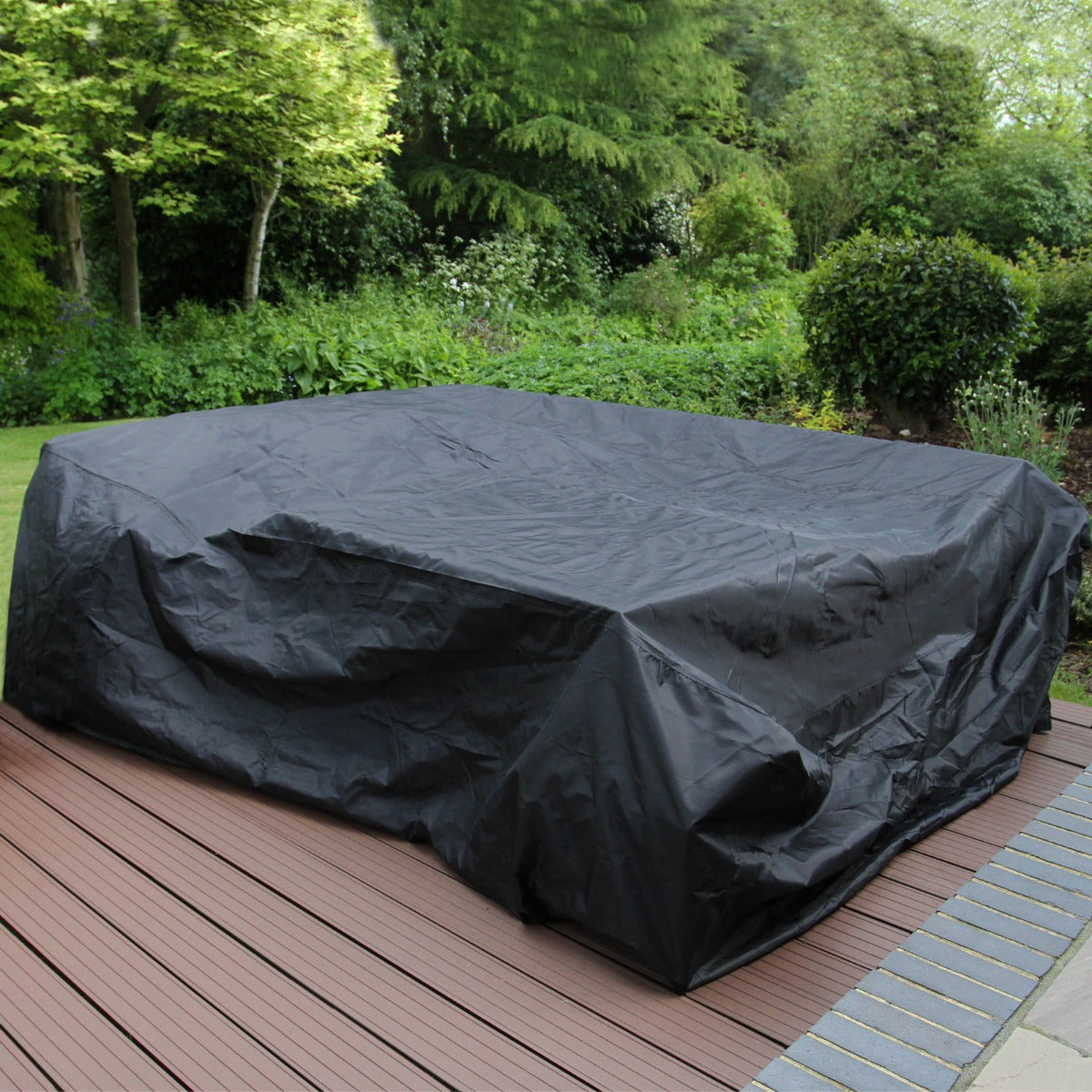 Grijze polyrattan hoekmeubelset met een beschermhoes, geplaatst op een houten terras omringd door groene planten en bomen