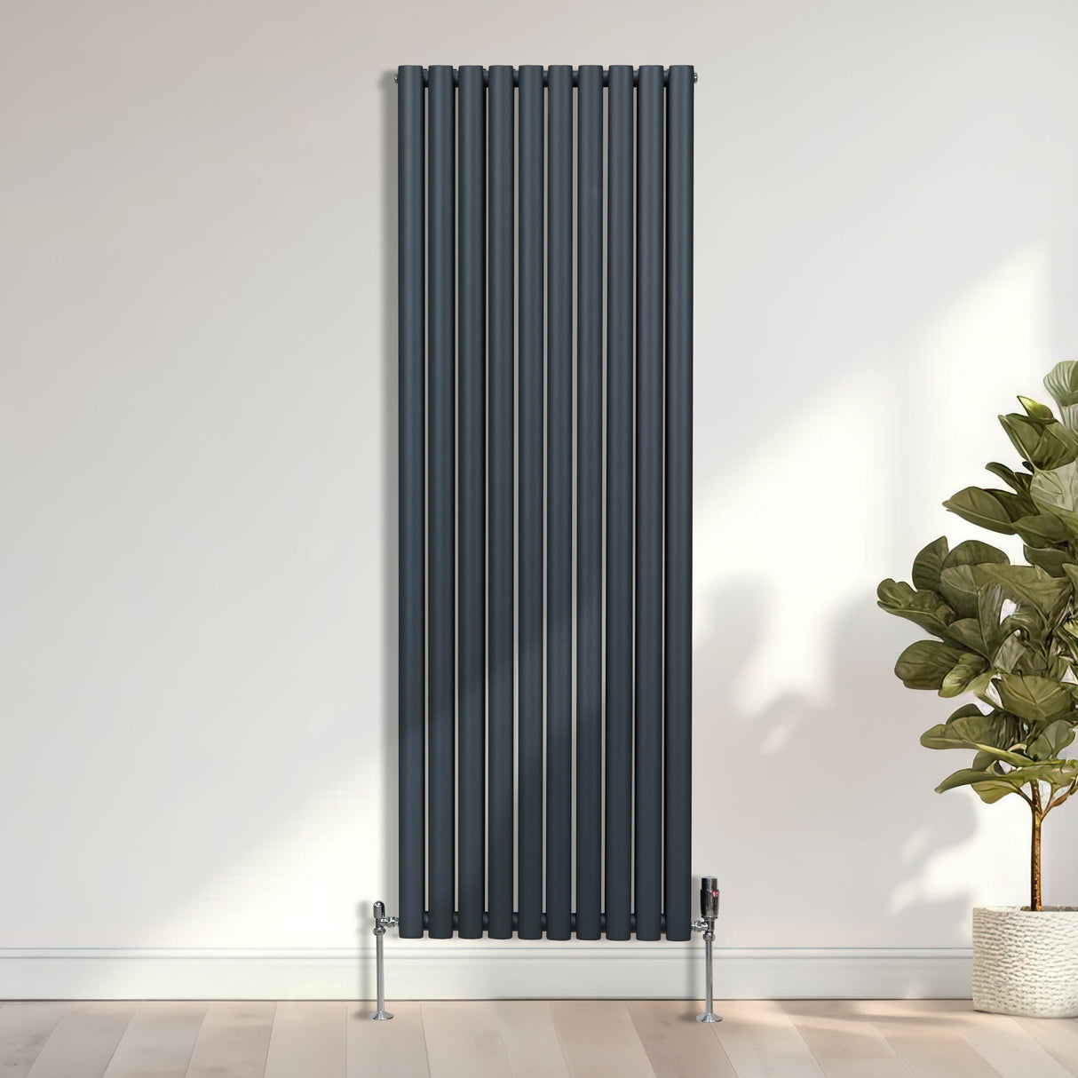 Moderne, verticale radiator in donkergrijs met chroom thermostatische kranen, geplaatst tegen een lichte muur met een plantenpot ernaast