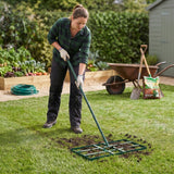 Groene 100 cm gazon leveler met lange steel, gebruikt door een vrouw in een tuin met groenten en een kruiwagen op de achtergrond