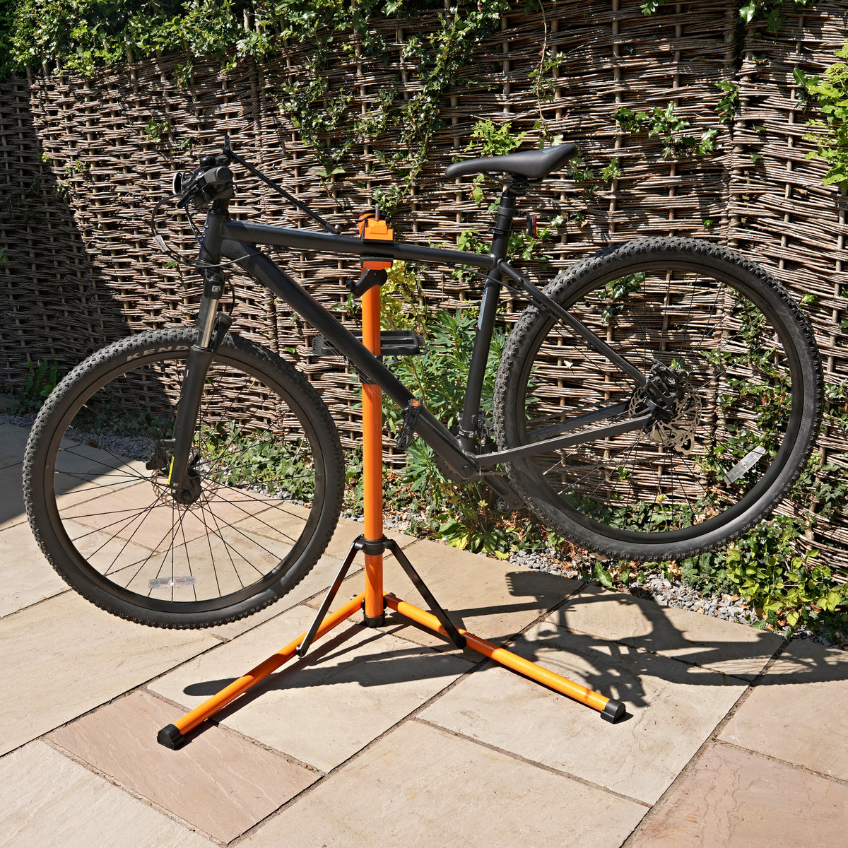 Zwarte fiets op een stevige oranje montagestandaard, met een natuurlijke achtergrond van een houten schutting en groene planten