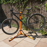 Zwarte fiets op een stevige oranje montagestandaard, met een natuurlijke achtergrond van een houten schutting en groene planten