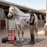 Een vrouw in een rijbroek en laarzen gebruikt een draadloze tondeuse op een grijs paard naast een paardendouche in een stal