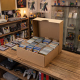 Een kartonnen doos gevuld met gesorteerde verzamelkaarten, geplaatst op een houten tafel in een retro gamewinkel met consoles op de achtergrond