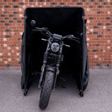Zwarte motorfiets met een robuuste band, gedeeltelijk verborgen in een zwarte hoes tegen een bakstenen muur