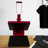 Rode hittepers met een zwart drukvlak van 38x38 cm, naast een zwarte en grijze T-shirt, op een houten tafel