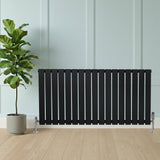 Zwart designradiator met vlakke panelen en haakse thermostaatkraan, geplaatst tegen een lichte wand met een groene plant ernaast