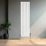 Witte designradiator met vlakke panelen en een haakse thermostaatkraan, geplaatst tegen een lichte muur met houten vloer en plant