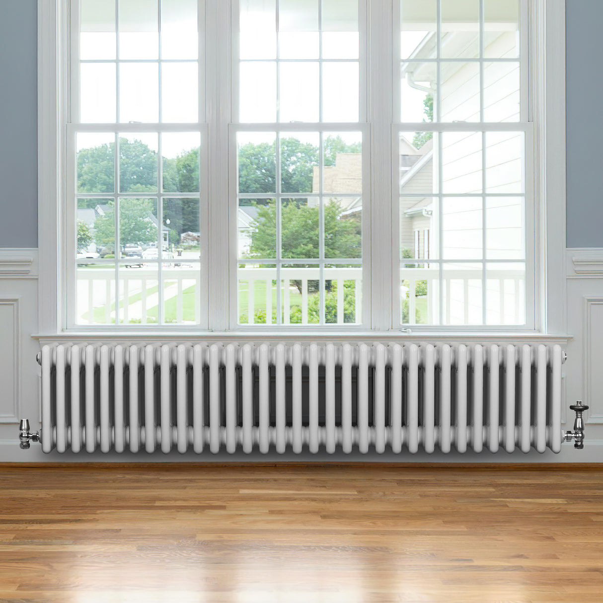 Traditionele witte 4-kolom radiator van 300 x 1505 mm met haakse thermostaatkraan, geplaatst onder een groot raam op houten vloer