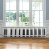 Traditionele witte 4-kolom radiator van 300 x 1505 mm met haakse thermostaatkraan, geplaatst onder een groot raam op houten vloer
