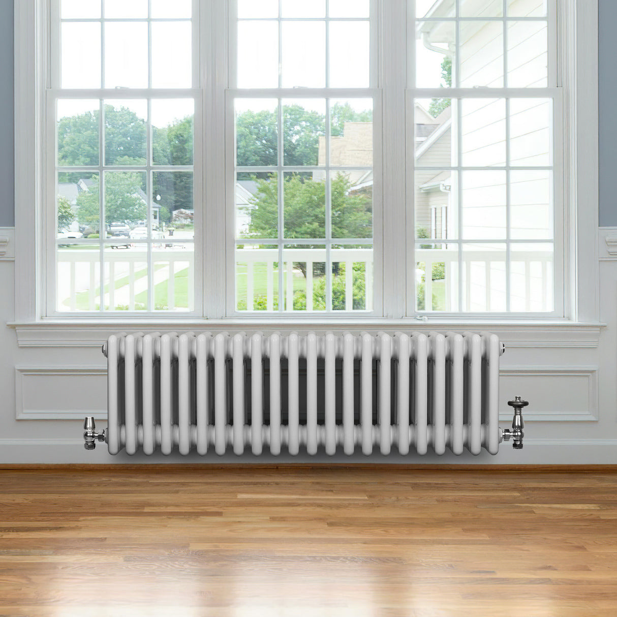 Traditionele witte 4-kolom radiator van 300 x 1010 mm, geplaatst onder een groot raam met uitzicht op een groene tuin en houten vloer