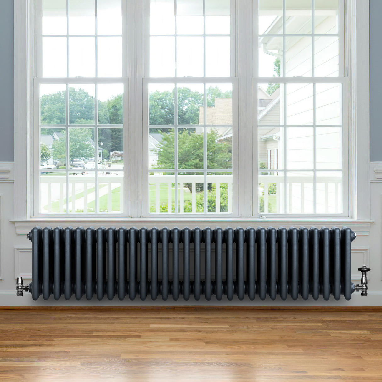 Traditionele 4-kolom radiator in antraciet grijs, met een glanzende afwerking, geplaatst onder een groot raam met houten vloer