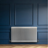 Traditionele witte 4-kolom radiator van 600 x 1010 mm tegen een donkerblauwe muur, met een strakke, glanzende afwerking en metalen aansluitingen