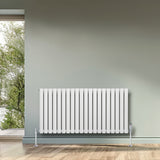 Ovale witte designradiator met één kolom, 600 mm x 1200 mm, geplaatst tegen een groene muur met houten vloer en natuurlijk licht
