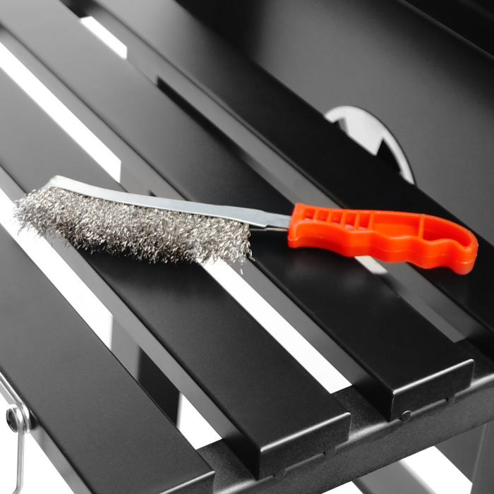 Roestvrijstalen borstel met een oranje handvat, ligt op een zwarte metalen ondergrond, ideaal voor het reinigen van grillroosters