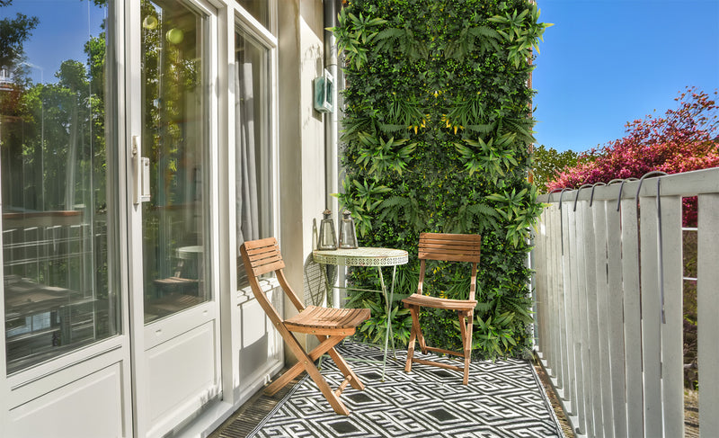 Een balkon met een groene wand van kunstplanten, een ronde tafel met twee houten stoelen en een zwart-wit tegelpatroon op de vloer