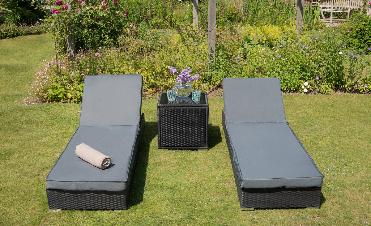 Twee comfortabele ligstoelen met grijze kussens en een zwart wicker tafeltje met bloemen in een groene tuin vol kleurige bloemen