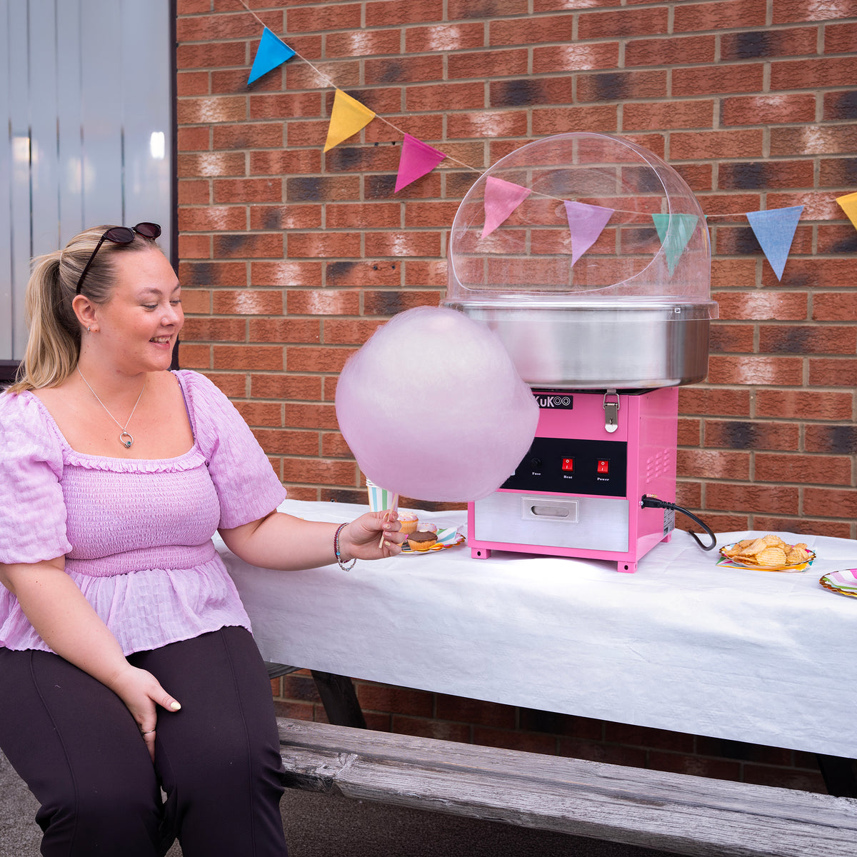 Een vrouw in een paarse blouse zit naast een roze suikerspinmachine, terwijl ze een grote, luchtige suikerspin vasthoudt