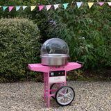 Felgekleurde suikerspinmachine in het roze met een transparante kap, geplaatst op een wagentje met wielen, omringd door groene struiken